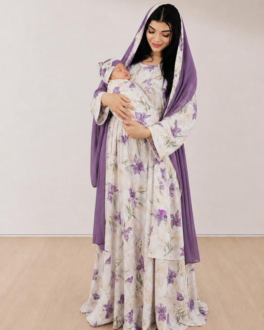 A woman wearing a long white dress with a purple floral print and a matching purple draped head covering, holding a swaddled baby. She is standing in a minimalist room with a light beige wall and light wood floor.